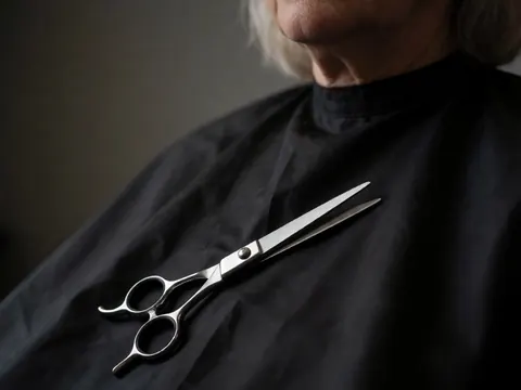 Des ciseaux métalliques posés sur une cape noire près du visage d'une personne aux cheveux clairs.