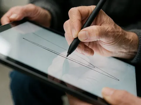Une main utilise un stylet noir pour tracer des lignes sur l'écran tactile d'une tablette, illustrant le travail numérique et la création assistée par technologie.