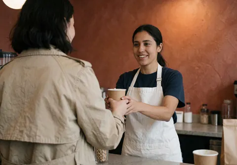 Une employée souriante remet une boisson à une cliente derrière le comptoir d'un café.