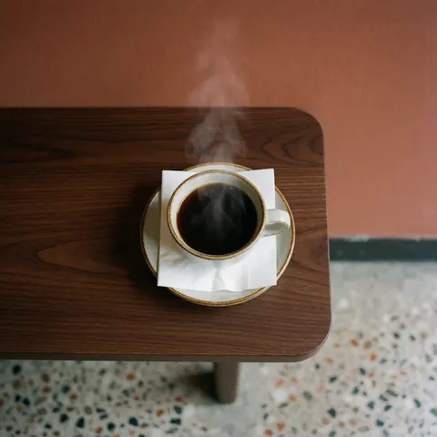 Une tasse de café noir fumant repose sur une serviette blanche, elle-même posée sur une table en bois sombre. La vapeur s'élève, évoquant la chaleur et le confort d'une boisson fraîche.