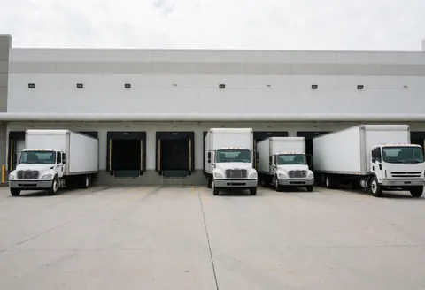 Plusieurs camions de livraison sont garés face aux quais de chargement d'un grand entrepôt logistique industriel.