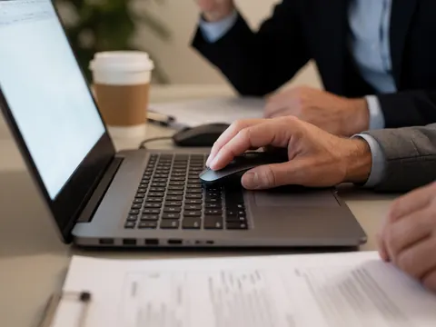 Un homme en costume utilise une souris sur un ordinateur portable posé sur un bureau avec des documents et une tasse de café.