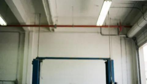 Vue du plafond d'un garage montrant des tuyaux, des luminaires fluorescents et une structure métallique industrielle en arrière-plan.