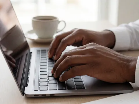 Gros plan des mains d'une personne tapant sur le clavier d'un ordinateur portable, accompagné d'une tasse de café posée sur la table.