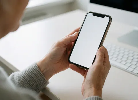 Un homme d'âge mûr tient un smartphone blanc devant un ordinateur sur un bureau clair, illustrant le télétravail ou l'utilisation professionnelle en intérieur.