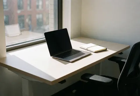 Bureau en bois avec ordinateur portable et cahier ouvert près d'une fenêtre laissant entrer la lumière naturelle.