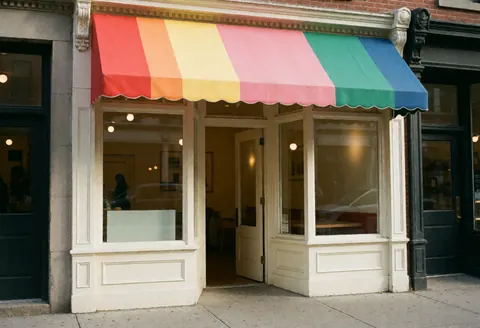 Vue extérieure d'une devanture de commerce affichant un auvent multicolore en forme d'arc-en-ciel, une fenêtre claire et une porte ouverte donnant sur la rue.