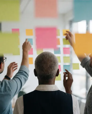 Trois personnes placent des notes adhésives colorées sur une paroi blanche lors d'une réunion professionnelle.