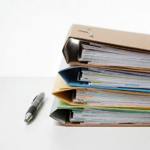 Une pile de dossiers contenant des documents repose sur une table blanche avec un stylo posé à côté.