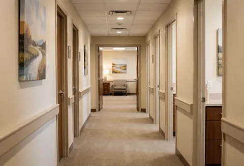 Intérieur d'un couloir de clinique aux murs beiges, portes de chambres et tableaux de paysages accrochés.
