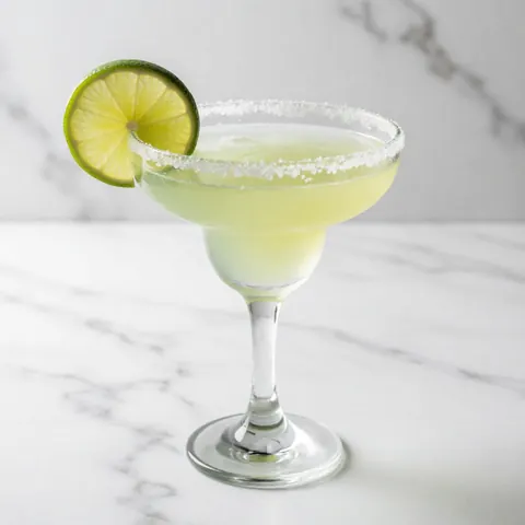 Un cocktail margarita avec du sel sur le rebord et une tranche de citron vert posé sur une surface en marbre blanc.