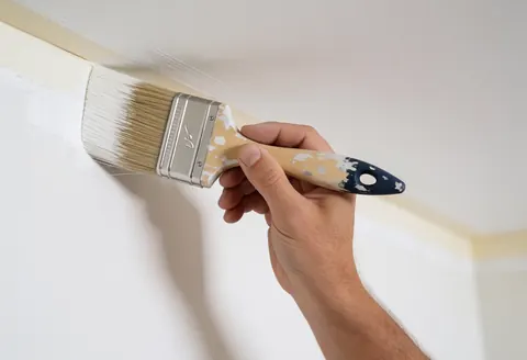Une main tenant un pinceau applique de la peinture blanche sur un mur intérieur près d'un angle, dans une vue rapprochée.