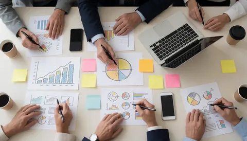 Des mains dessinant des graphiques et diagrammes sur une table avec un ordinateur portable, des téléphones et des post-it lors d'une réunion professionnelle.