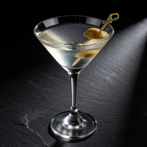 Un cocktail martini garni d'une olive est servi dans un verre à cocktail posé sur une table en pierre sombre, éclairé par une lumière dramatique.