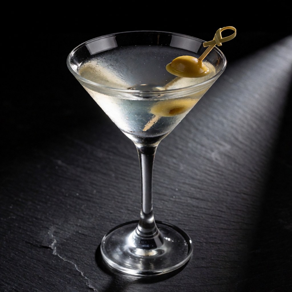 Un cocktail martini garni d'une olive est servi dans un verre à cocktail posé sur une table en pierre sombre, éclairé par une lumière dramatique.