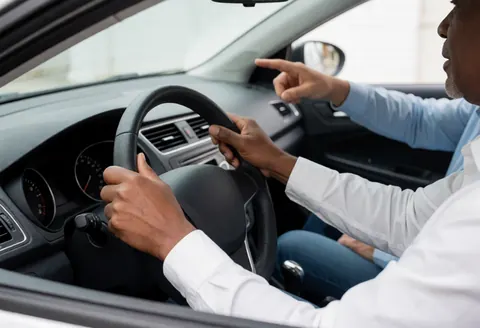 Un instructeur de conduite donne des instructions à un élève pendant qu'il conduit une voiture, avec une main sur le volant et l'autre pointant.