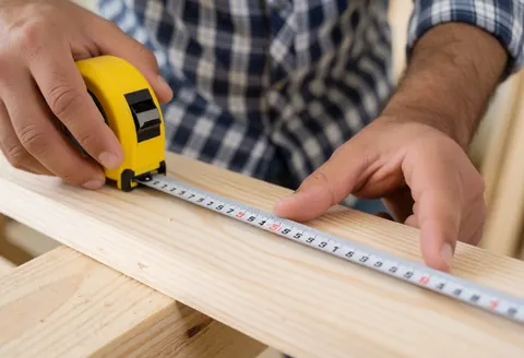 Un menuisier mesure une planche en bois à l'aide d'un mètre ruban jaune dans son atelier de travail.