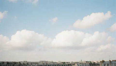 Vue d'un ciel bleu parsemé de nuages blancs au-dessus des toits d'une ville européenne, offrant un fond urbain minimaliste et épuré.