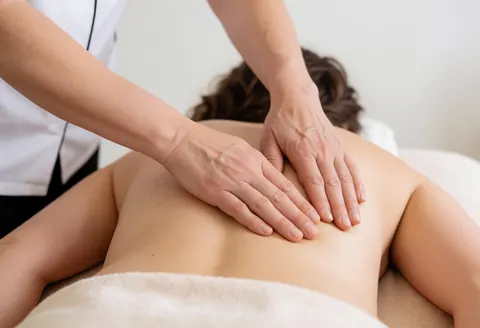 Une praticienne réalise un massage sur le dos d'une cliente allongée sur une table recouverte de serviettes blanches.
