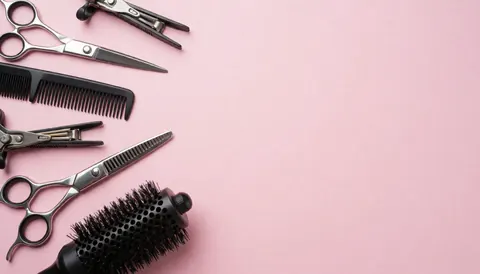 Ciseaux, peigne et brosse disposés sur un arrière-plan rose pastel uni, évoquant l'univers professionnel du salon de beauté.