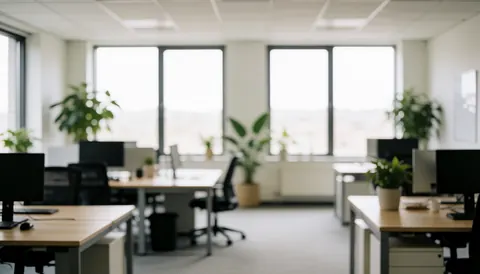 Vue floue d'un bureau moderne comprenant des bureaux, des ordinateurs et des plantes, créant un arrière-plan neutre et professionnel pour du texte.