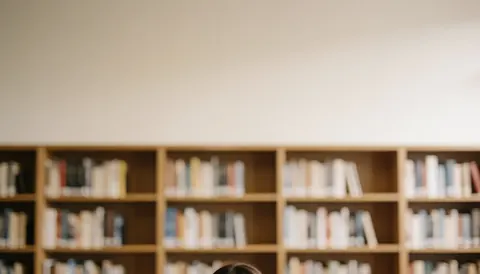 Vue arrière d'une personne debout face à des rayonnages remplis de livres dans une bibliothèque baignée d'une lumière chaleureuse et apaisante.