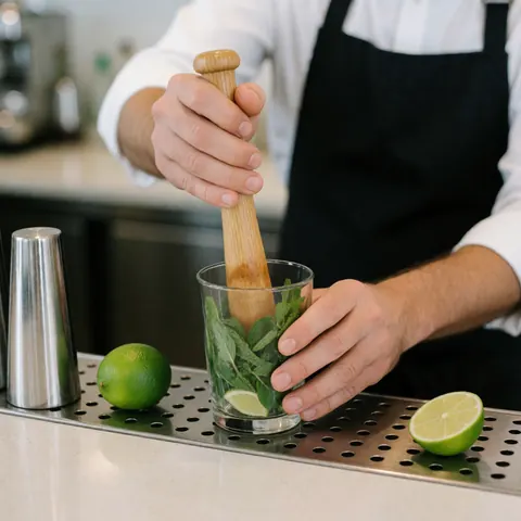 Un bartender utilise un pilon en bois pour écraser de la menthe et du citron vert dans un verre sur un comptoir métallique.