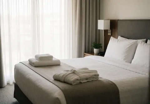 Lit d'hôtel avec draps blancs, serviettes et peignoir plié près d'une fenêtre avec rideaux.