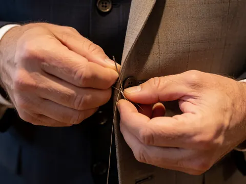 Gros plan des mains d'un homme en train de coudre un bouton sur une veste à carreaux, illustrant le travail artisanal et la couture.