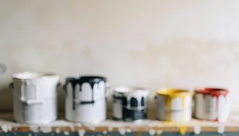 Plusieurs seaux de peinture aux couleurs variées posés sur une étagère sale avec des gouttes, devant un mur blanc taché servant de fond.