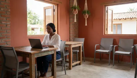Une femme utilise un ordinateur portable sur une table en bois dans un espace de coworking lumineux aux murs terracotta, avec des plantes et du macramé.