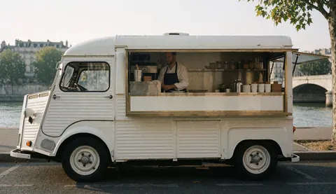 Un food truck blanc est garé au bord de l'eau avec un homme préparant des boissons derrière le comptoir.