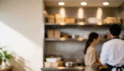 Deux personnes travaillent dans une cuisine professionnelle équipée d'étagères et baignée d'une lumière chaude.