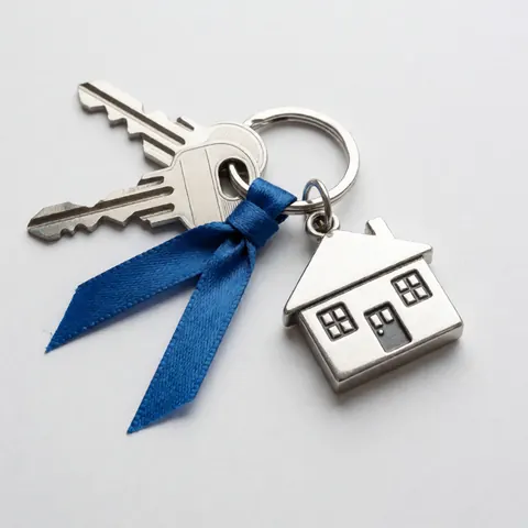 Des clés métalliques avec un porte-clés en forme de maison et un ruban bleu disposés sur un fond blanc uni, vue de dessus.