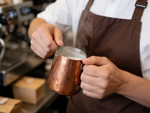 Un barista utilise une carafe en cuivre pour chauffer du lait dans un établissement de café, mettant en valeur le matériel professionnel et la préparation minutieuse des boissons.