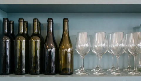 Des bouteilles de vin et des verres sont disposés sur une étagère en bois dans un bar, créant un fond neutre pour du texte.