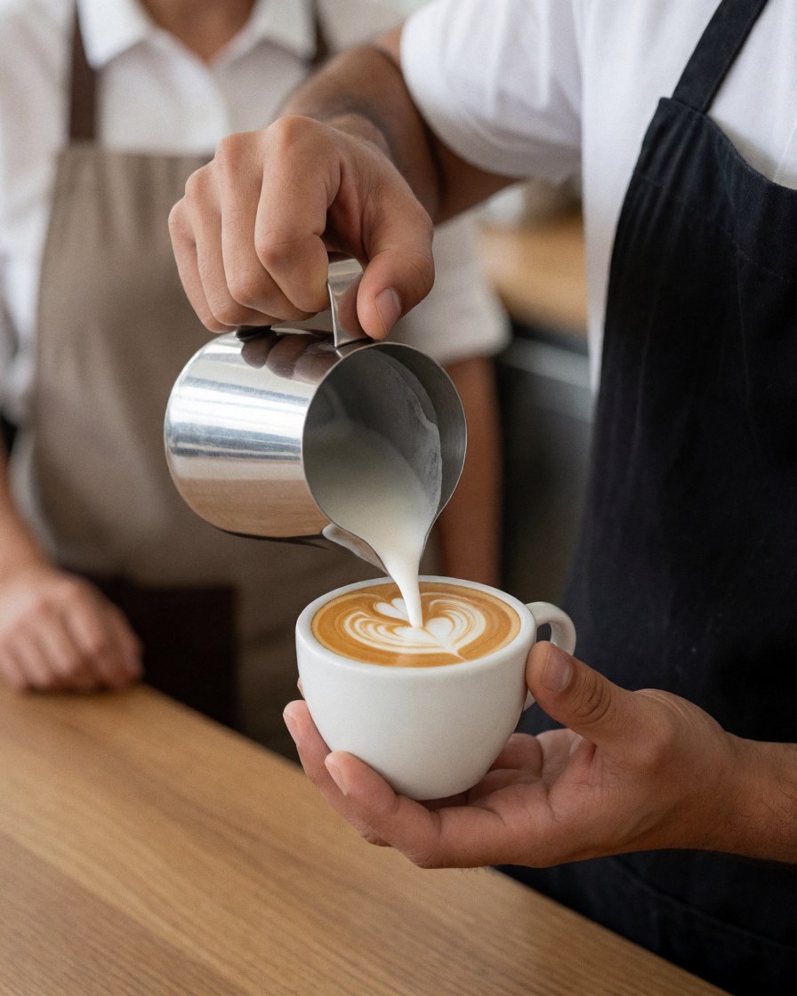 Un barista en tablier noir verse du lait dans une tasse de café sur un comptoir en bois pour réaliser un motif artistique.
