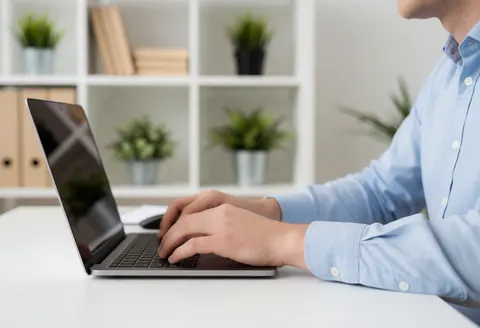 Un homme adulte portant une chemise bleue travaille sur un ordinateur portable placé sur une table blanche dans un bureau moderne avec étagères et plantes.
