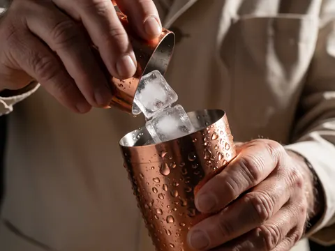 Main d'un barman versant des cubes de glace dans un shaker en cuivre parsemé de gouttelettes d'eau.