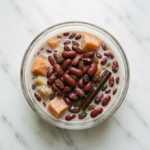 Un verre contenant des haricots doux au lait de coco posé sur une surface en marbre blanc, garni de fruits, de raisins secs et de cannelle.