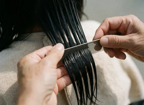 Détail d'une coupe de cheveux avec un peigne fin, cheveux humides et brillants, serviette beige, mains travaillant avec précision, éclairage naturel, détail professionnel.
