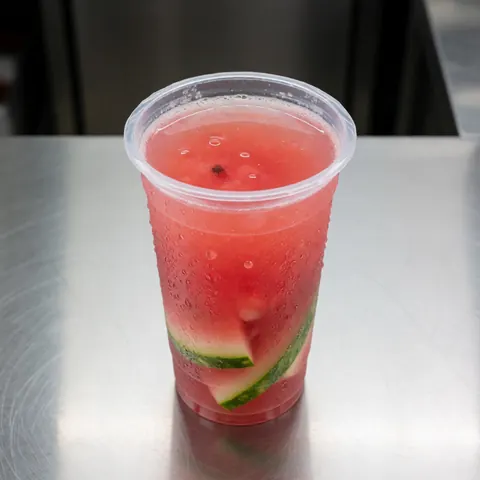 Un verre en plastique rempli d'eau de pastèque avec des morceaux de fruit, posé sur le comptoir d'un food truck, offrant une boisson rafraîchissante et appétissante.