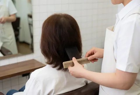 Un coiffeur professionnel coupe les cheveux bruns d'une cliente dans un salon de coiffure moderne avec miroir et outils visibles.