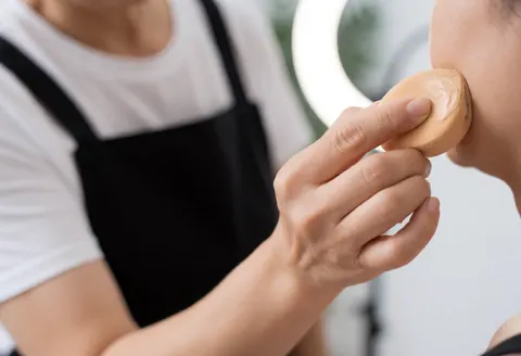 Une maquilleuse professionnelle applique de la base sur le profil d'une cliente à l'aide d'une éponge humide. Éclairage par ring light, fond flou dans un salon de beauté.