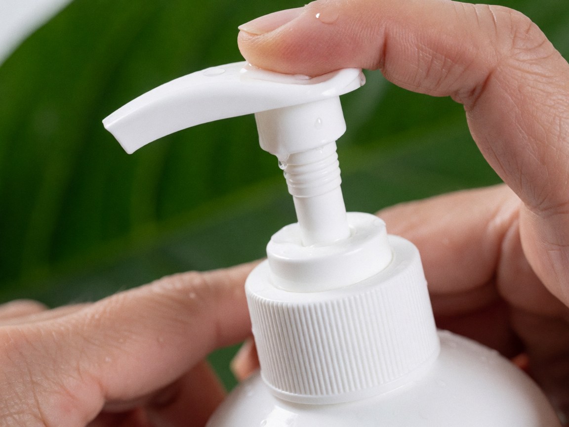 Gros plan d'une pompe blanche de shampooing avec des gouttes d'eau, tenue par des mains, sur un fond de feuille verte, évoquant fraîcheur et soin naturel des cheveux.