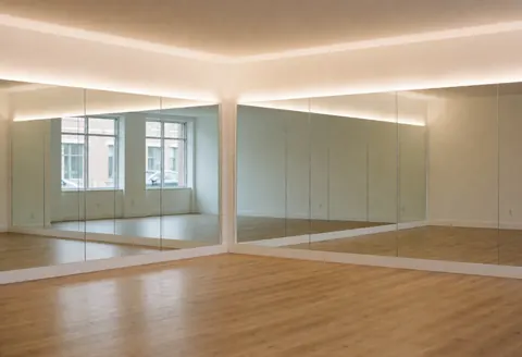 Studio de yoga vide avec miroirs, sol en bois clair et lumière chaude, idéal pour cours de yoga ou mobilité, ambiance calme et spacieuse.