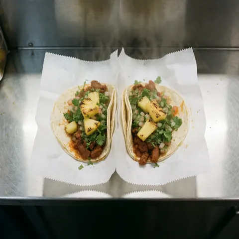 Vue rapprochée de tacos al pastor garnis d'ananas, de coriandre et d'oignon sur un comptoir en bois.