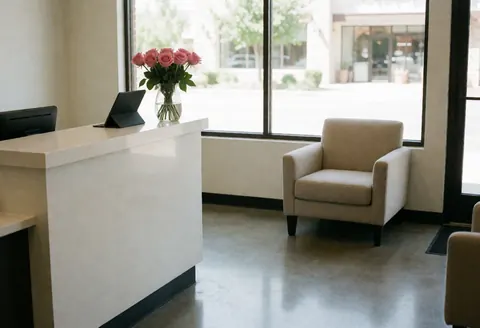 Intérieur d'une réception de salon de coiffure moderne avec une chaise beige et rose, une tablette, une fenêtre laissant entrer la lumière naturelle, un sol en béton poli, ambiance propre et accueillante.