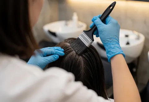 Un coiffeur portant des gants bleus applique une teinte sombre au pinceau sur les racines d'un client dans un salon avec lavabos blancs et murs en béton.