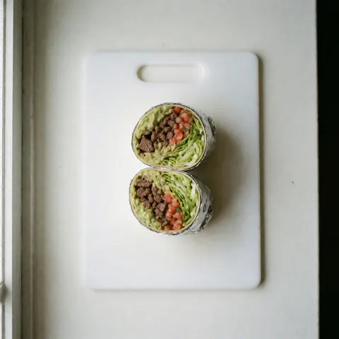 Un burrito tranché en deux révélant sa garniture de viande, guacamole, laitue et pico de gallo posé sur une planche blanche éclairée par la lumière naturelle.